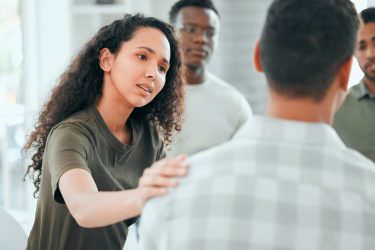 Counseling, woman with hand on man shoulder for comfort and in support center with members. Mental health or depression, group therapy or rehabilitation and people sitting together for recovery.